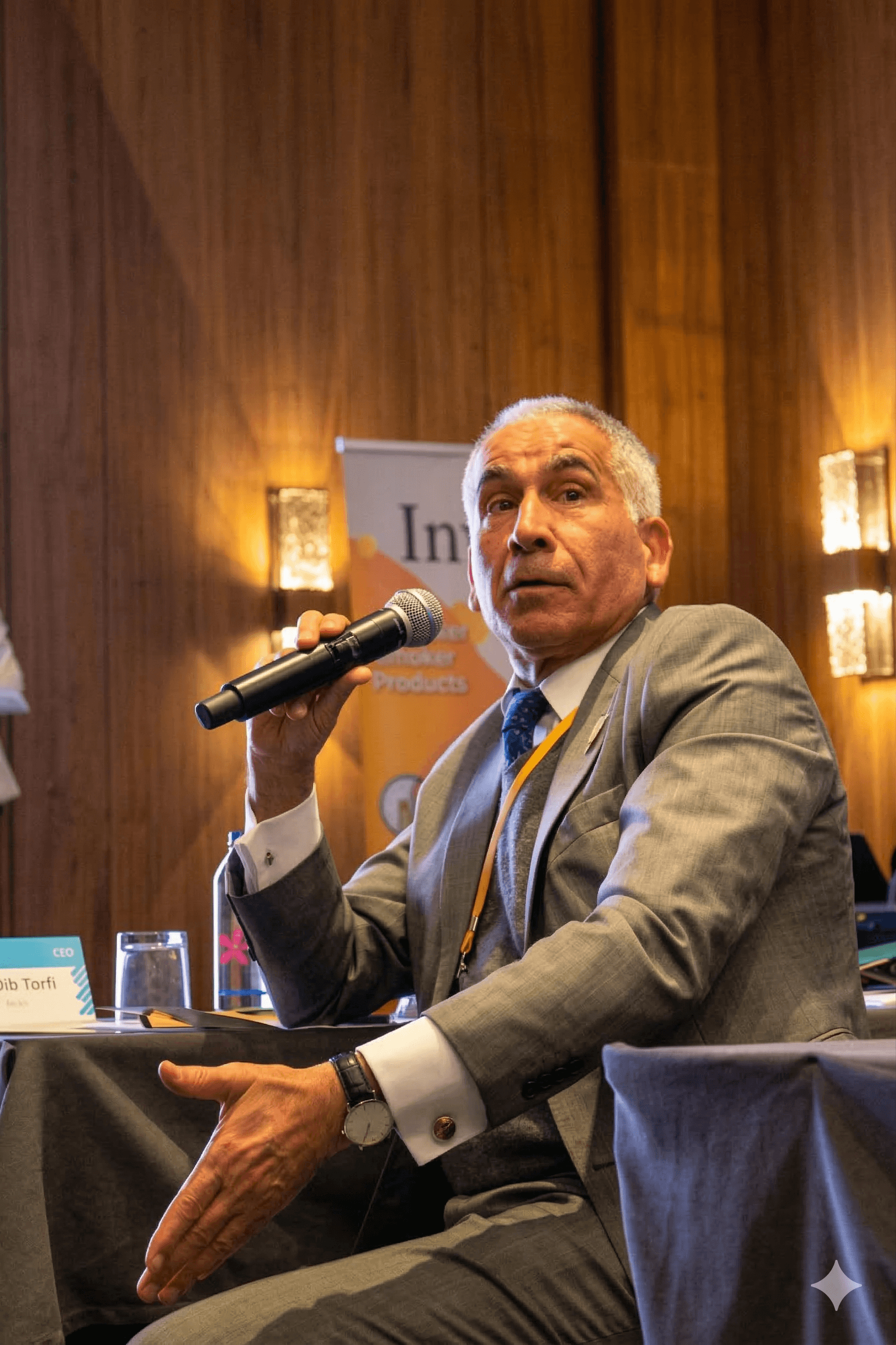 Man in a grey suit gestures while speaking into a microphone at a conference.